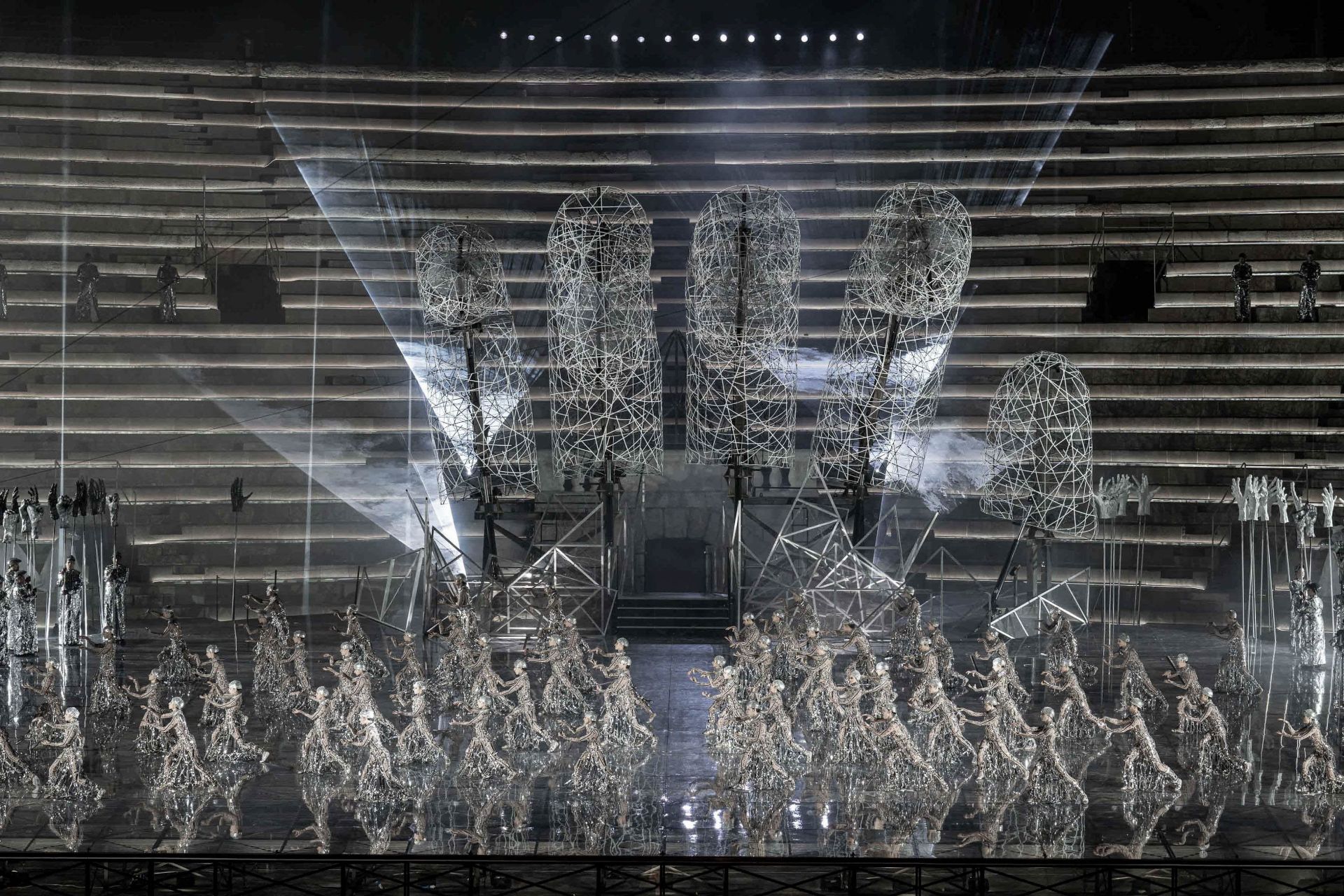 Act II. The stage is again dominated by the large hand, illuminated by beams of white light and surrounded by dense clouds of smoke. At the back of the stage, the spears with white hands can be seen to the right while the spears with the black hands are to the left. They are all made of papier-mâché. In front of the large hand, male and female dancers occupy the entire stage. They are standing and facing the right with both their right leg and their arms extending forwards. They are wearing tight beige colored suits decorated with designs of black hieroglyphs. On top of their suits, they are wearing sparkling silver netting accented with crystals with silver caps on their heads.