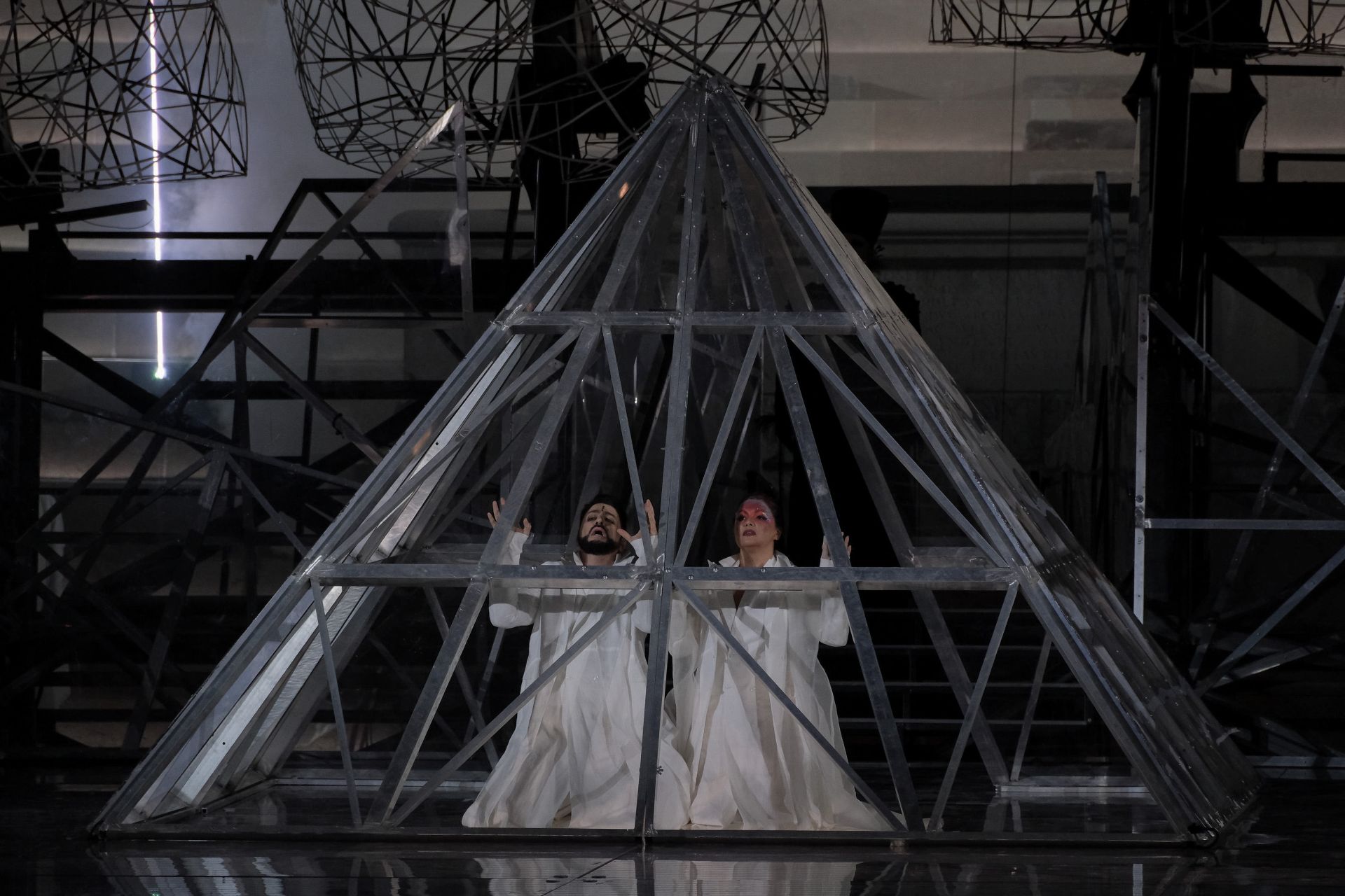 Act IV. Radamès and Aida are on their knees side by side within an iron structure in the form of a pyramid that recalls the one outside the Louvre. Both are dressed in white and hold their hands up in front of them. Aida is wearing light red and purple makeup on her forehead covered with multicolored glitter, while Radamès, has a small black line at the center of his.