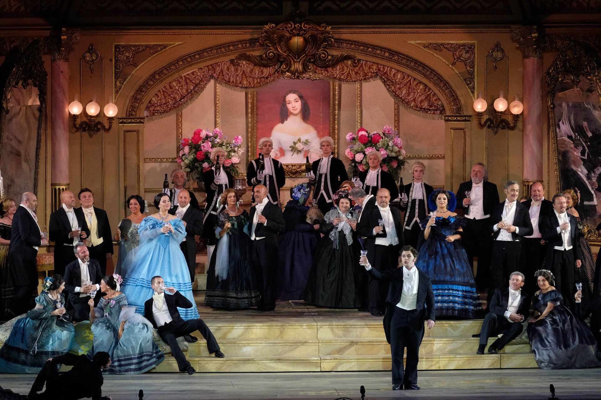 First act. Alfredo, Violetta and some of the guests at the party held at Violetta’s house. The picture shows the sitting room in Violetta’s house. The back wall is gold in color and richly decorated in the style of the 19th century, with stuccoes, arches and mirrors. At the center of the wall hangs a large head and shoulders portrait of Violetta. In the painting, she is wearing an elegant white strapless gown with a rose pinned to the center of her breast. At both sides, there are two wall lights and two large vases filled with flowers standing on a raised surface. In front of the portrait, waiters in livery are standing on steps and holding bottles of champagne. Further toward the front, there are other stairs where some of the guests are sitting with glasses in hand. At the center of the scene, Alfredo is in the act of raising his own glass.