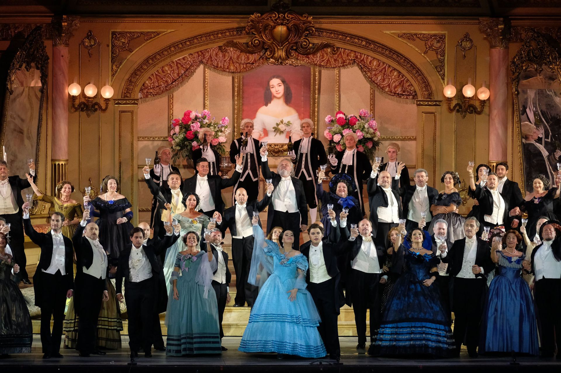 Violetta, Alfredo and some of Violetta’s friends. In the first act, Violetta is wearing an elegant light blue gown. The bodice leaves her shoulders bare, while the skirt billows out from the waist and is floor length with two ruffles at the bottom. The gown is overlaid with light blue dotted tulle that ends with blue rings. She is also wearing a precious parure of jewelry, with matching earrings and necklace, along with a tulle shawl that partially covers her shoulders and arms. Her hair is brown and pulled back. Alfredo is wearing a black tailcoat, with pants and a short coat that ends at the waist with two long tails falling to his knees in back. Beneath his coat, he is wearing a white vest and white shirt, as well as a white scarf tied around his neck. He has short, brown hair. The female guests at the party are all wearing elegant, puffy gowns in shades of light blue and blue. The men are wearing tailcoats, like Alfredo. The waiters are wearing black livery with grey trim. They are also wearing white wigs with curls at ear level.