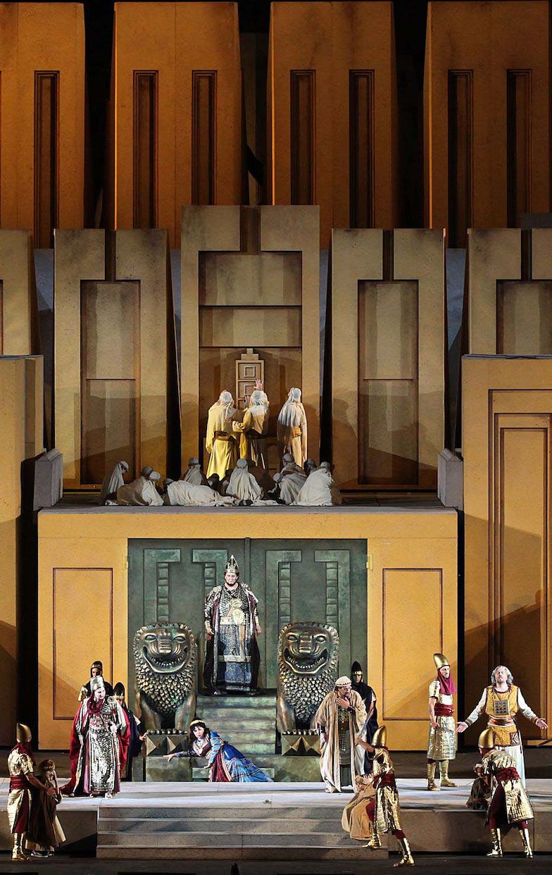 Nabucco, Fenena, Abigaille, Part I – Nabucco is standing at the center of the stage, Fenena is half lying at his feet and  Abigalle is standing to the left. Nabucco is wearing a light blue, Bordeaux and ocher robe with exquisite gold accents. On top of the robe, he is wearing a mantle with the same colors as the robe and it too is exquisitely decorated. He is wearing a gold headdress, similar to a helmet, that leaves his face uncovered. On his arms, he is wearing gold cuff bracelets. Fenena is wearing a long white dress with long sleeves. It is made of a variety of fabrics and colors with shades of light blue, gray and Bordeaux. Around her waist she is wearing a belt made of gold medallions. Her hair is long and dark brown. She is wearing a gold band on her head. Abigaille is wearing a long dress similar to armor with shades of gold and red. On top, she is wearing a floor length red mantle with gold accents. On her head she is wearing a gold headdress, similar to a helmet, that leaves her face uncovered. On her arms she is wearing gold cuff bracelets.