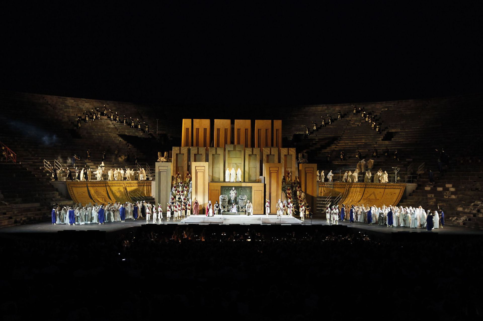 Part I – Behind the stage are the concentric tiers of seats that form the arena. The center of the stage is dominated by the temple of Jerusalem, built using seventeen enormous blocks in shades of orange and in the shape of rectangular parallelepipeds positioned horizontally. At the center of the structure is a dark green block with the same shape but positioned horizontally that encloses the entrance to the temple. In front of the door stand two silver statues of majestic lions. There is a flight of steps between the two statues. Nabucco is standing on the last stair while Fenena is half lying on the first. To the left of one of the statues stands Abigaille, while Ismaele is to the right of the other statue.  At both sides of the temple, large rectangular wall hangings in shades of orange descend from approximately the height of the tenth tier of seats towards the stage. Babylonian warriors, Hebrews, Levites and Hebrew virgins are standing around the temple and at both sides. 