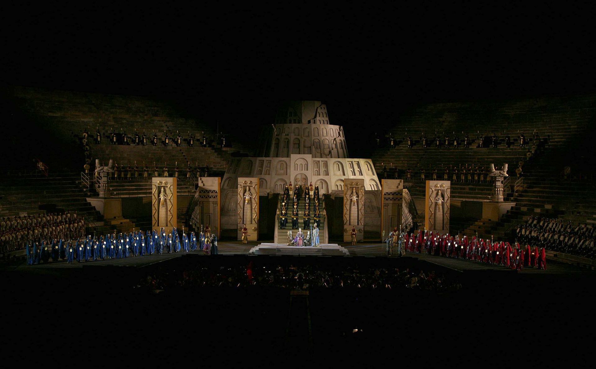 Part III – Just as in part II, the center of the stage is dominated by the royal palace of Babylon made with gray-ocher colored blocks and surmounted by a circular ziggurat that recalls the Tower of Babel in the famous painting by Pieter Bruegel. In front of the palace stand six enormous blocks in the shape of rectangular parallelepipeds that are the same color as the palace. Three stand to the left and three to the right, creating a space in the center occupied by a flight of steps. On the steps and at the sides of the stage soldiers, women of Babylon and the populace are standing. At the center of the stage, in front of the flight of steps, Abigaille is sitting on a gold throne without a backrest.  