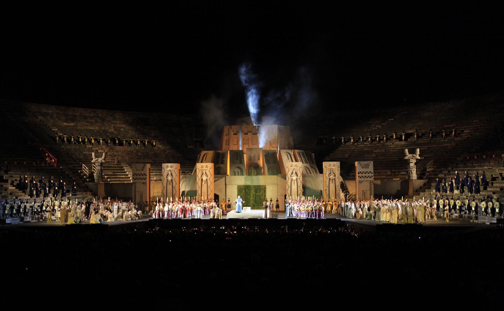 Part IV – The center of the stage is still dominated by the royal palace of Babylon. The blocks that form the top are now slightly set apart from one another and dense gray smoke is coming out of them from within. In front of the entrance, Nabucco is standing with his arms spread out and is surrounded by Zaccaria, Anna, Fenena, the High Priest of Baal, magi, Hebrews, guards and the people.