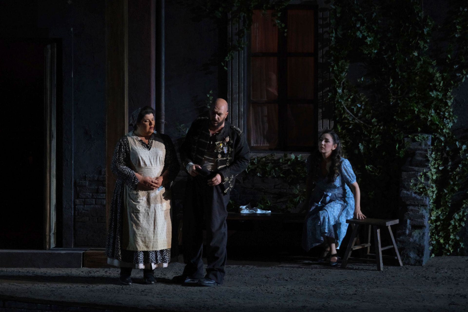 Giovanna and Rigoletto are standing next to each other.  Gilda is sitting to the right. Giovanna is wearing a blue dress with a floral design with white apron over the dress. On her head she is wearing a gray scarf that pulls her hair together. Rigoletto is wearing a short, dark blue military jacket with 7 rows of gold embroidery around the button. He has several medals pinned to his chest. Beneath the jacket, he is wearing a wrinkled gray flannel shirt. He is wearing dark blue pants and work shoes with a black beret on his head. Gilda is wearing a light blue and white dress with a floral design with the hemline at her knees. She is wearing black flat shoes. Her hair is down and pinned to her head.  Around her neck she is wearing a gold chain with a cross