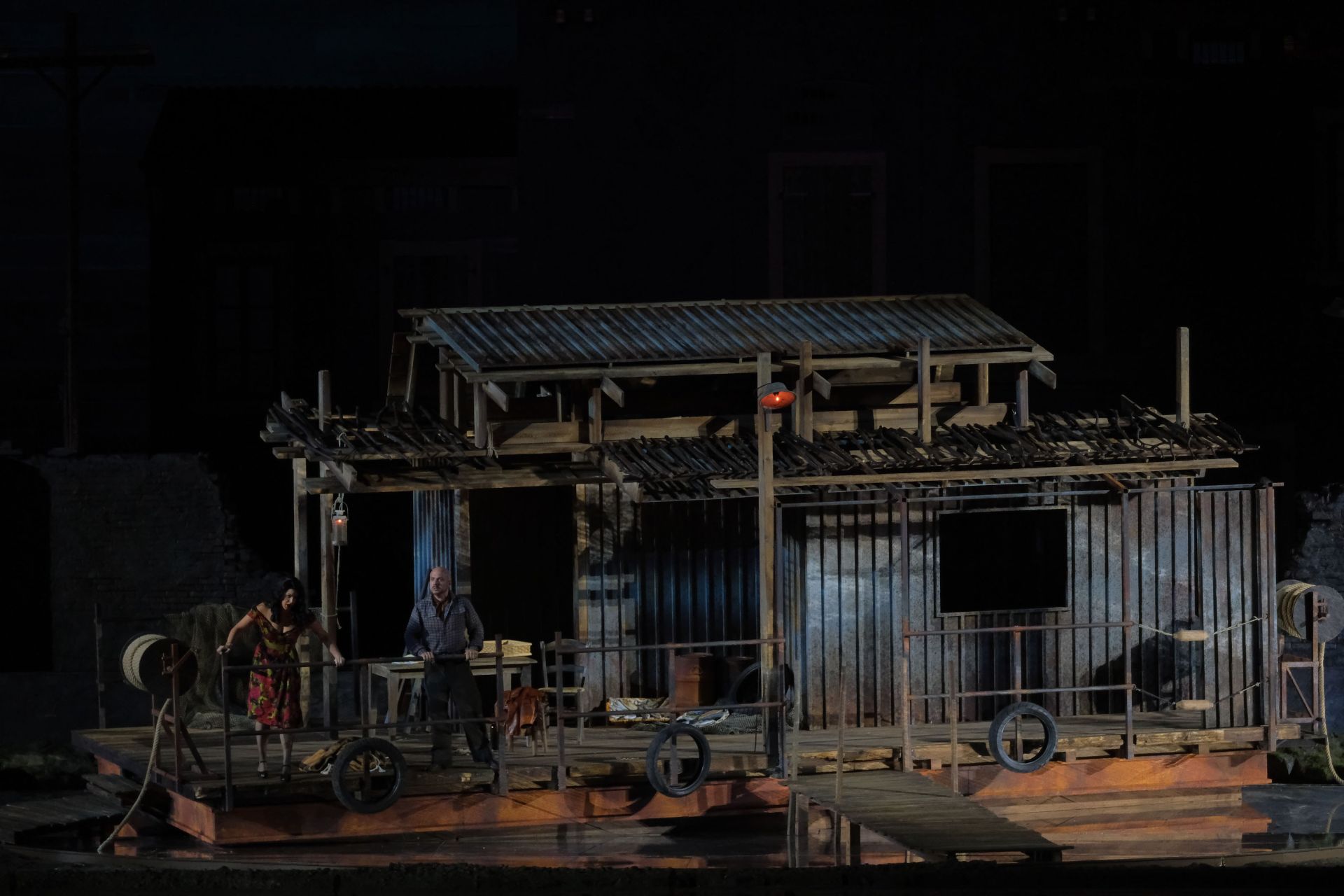 The image shows Sparafucile’s inn/raft: it is a flat bottomed boat, 13 meters wide and 7 meters long. A gangway connects it to to shore. The raft is made out of rusty sheet metal and wooden beams. A lamp hangs from one of them.   To the left a chair and table are on the deck of the raft. Along the outside three tires are tied to a metal railing, while there are two coils of rope on the sides. 